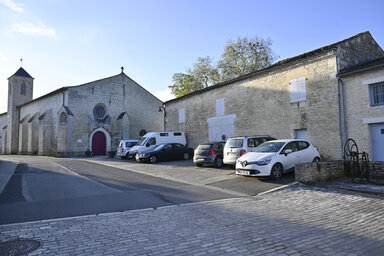Parvis pavé devant une église en pierre de taille avec clocher et bâtiments anciens environnants