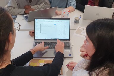 Femmes réunies autour d'ordinateurs portables lors d'un atelier collaboratif, l'une désignant une interface à sa voisine