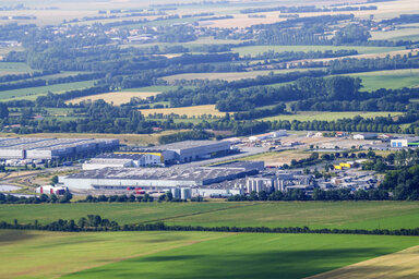Survol aérien des entrepôts et bâtiments industriels de la zone, entourés de champs agricoles et de boisements