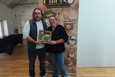 Deux personnes présentant la boîte de Circino 79 devant le kakémono du jeu « Le chasseur de trésors »